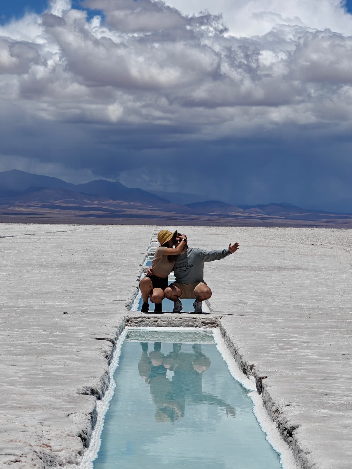 A couple crouching beside a turquoise salt pool at Salinas Grandes in Jujuy